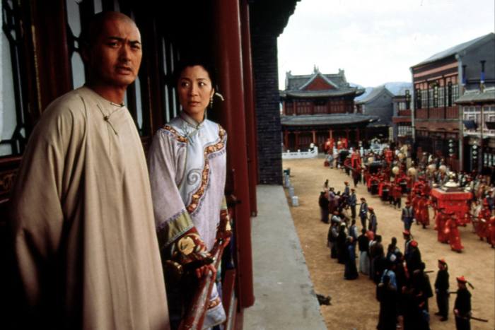 A man in a woman in period Chinese dress look stand on a balcony, above a street where a procession is taking place